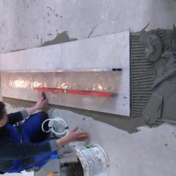 Rem-Bud Bartosz Żemła - Glazurnik w trakcie pracy: przyklejanie podłużnej płytki ściennej z użyciem poziomicy, widoczny klej na ścianie i narzędzia w wiadrze.