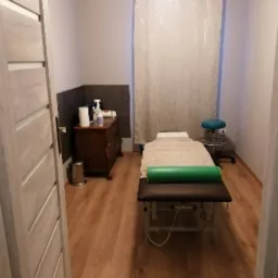 Jasne wnętrze gabinetu masażu z drewnianą podłogą, brązową komodą z akcesoriami, turkusowym stołkiem i profesjonalnym łóżkiem do masażu z zielonym wałkiem, widziane zza uchylonych drzwi.