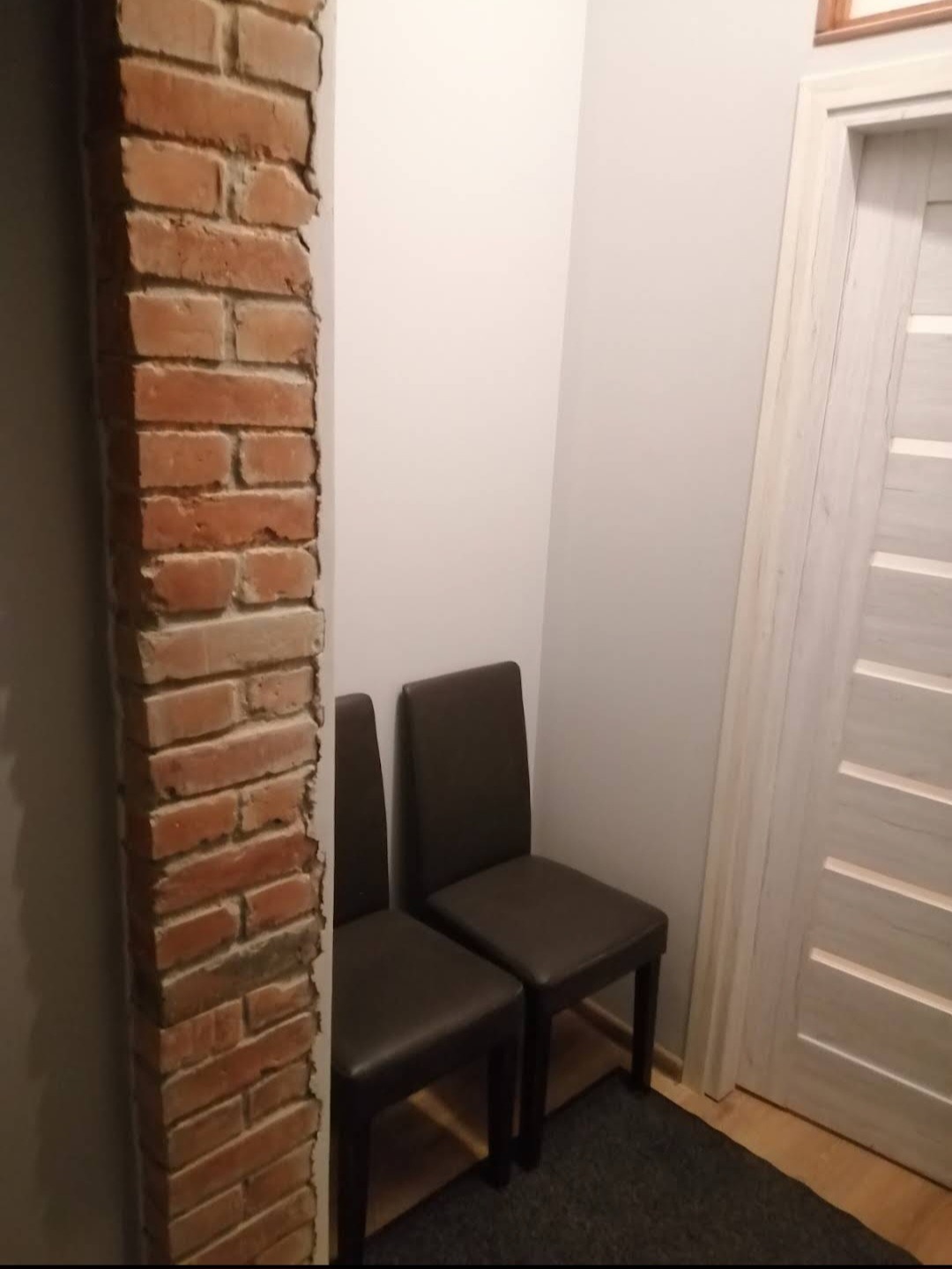 Dwa ciemne krzesła z ekoskóry stojące przy ścianie i drzwiach, obok fragmentu ściany z surowej cegły.