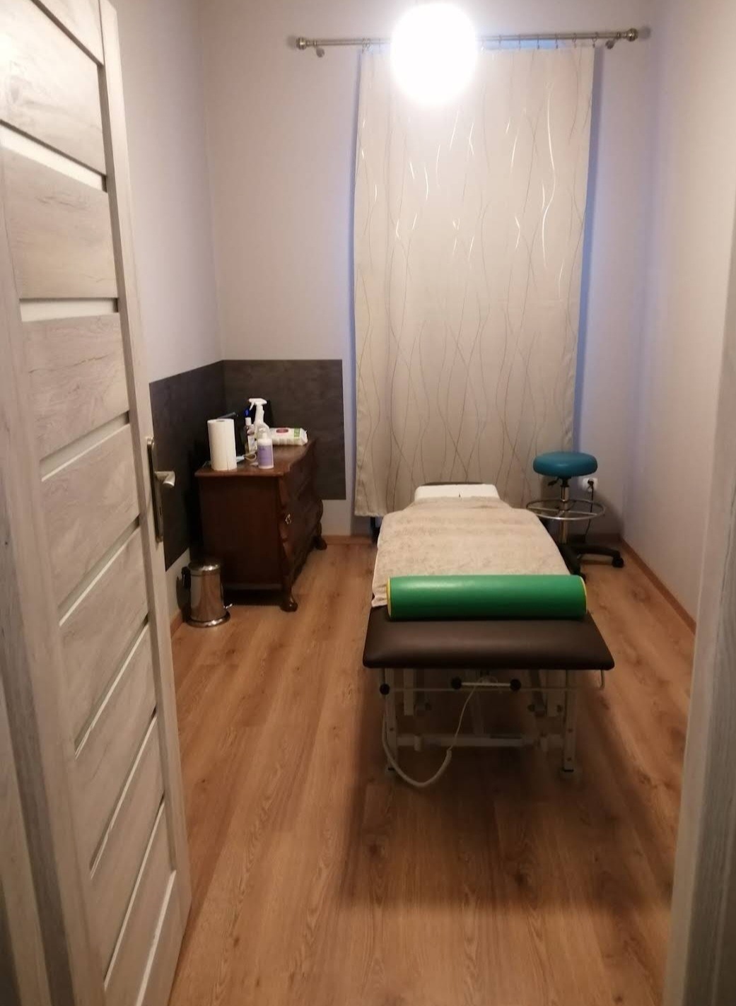 Jasne wnętrze gabinetu masażu z drewnianą podłogą, brązową komodą z akcesoriami, turkusowym stołkiem i profesjonalnym łóżkiem do masażu z zielonym wałkiem, widziane zza uchylonych drzwi.