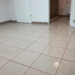 Wypolerowana, jasna podłoga z płytek w pustym pomieszczeniu biurowym z białymi drzwiami i częściowo widocznymi szafkami w tle.