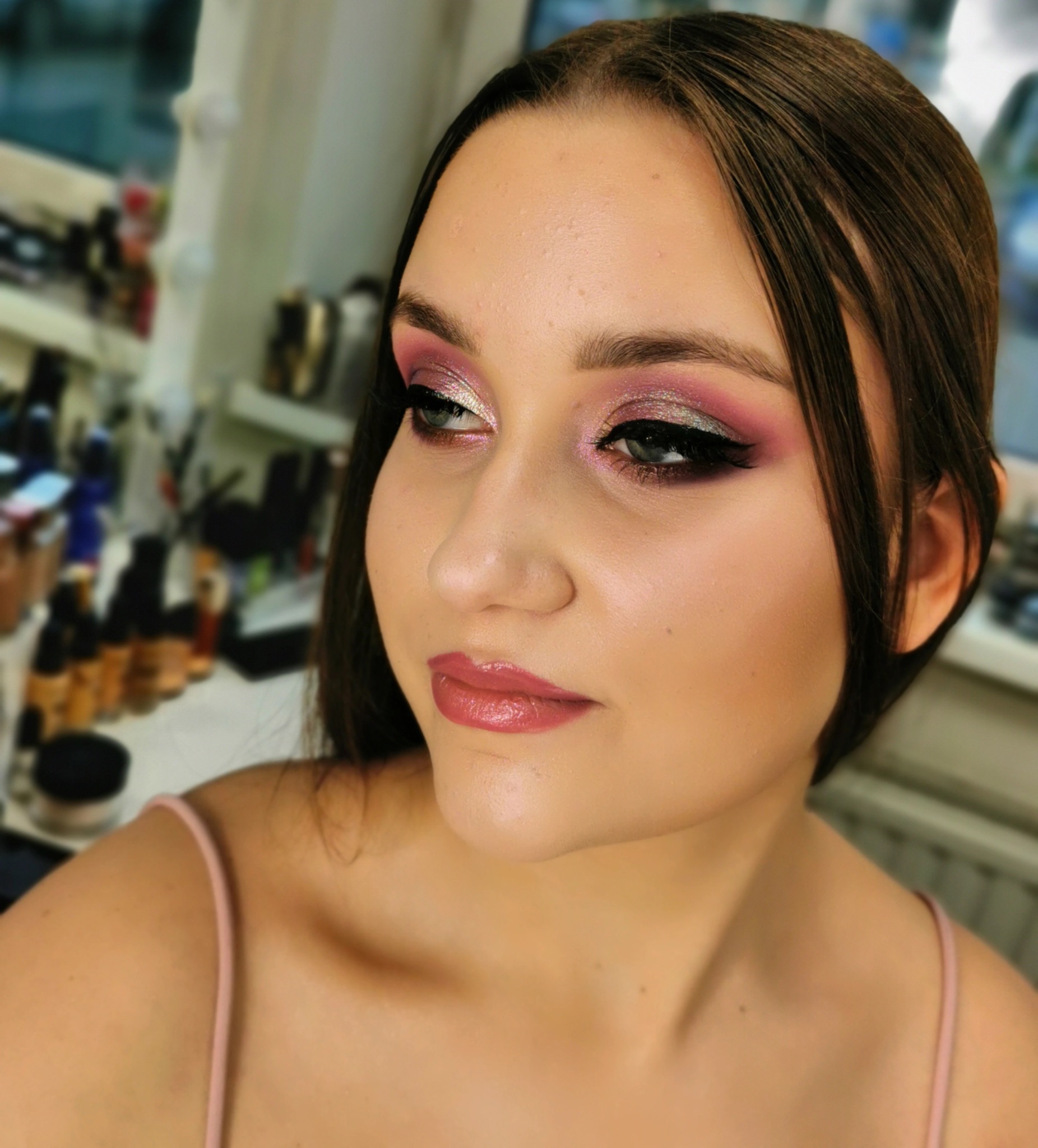 Portret kobiety z profesjonalnym makijażem: różowo-srebrne cienie, precyzyjna kreska eyelinerem, błyszczące usta, z widocznym tłem z kosmetykami.