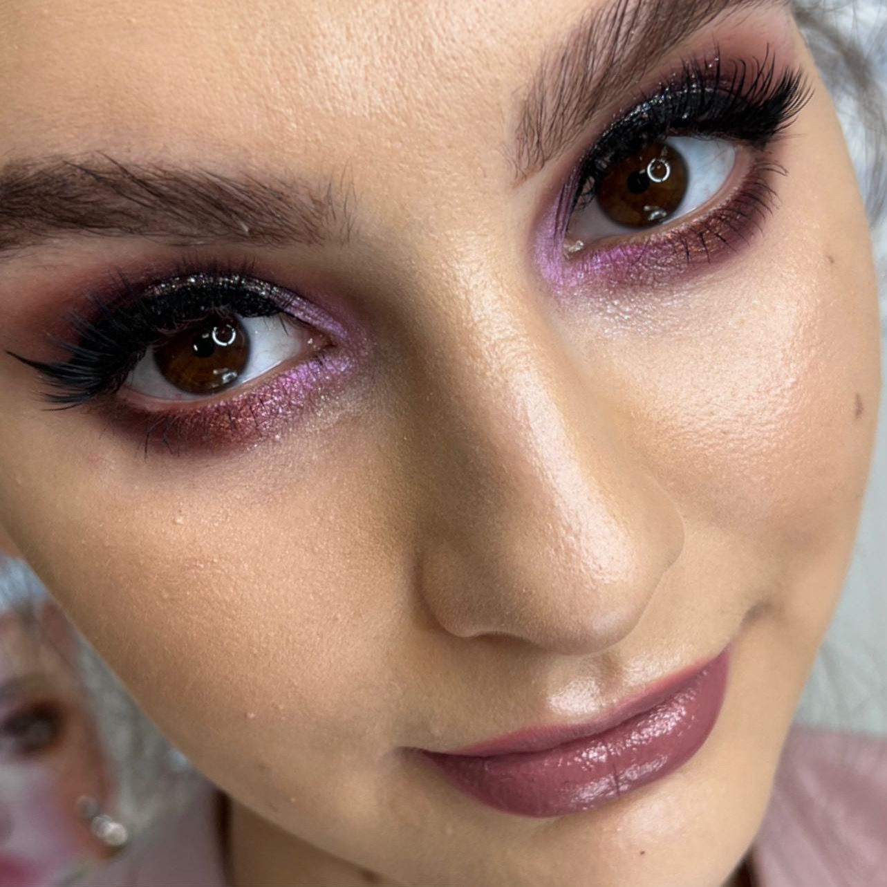 Portret kobiety z intensywnym makijażem oczu w odcieniach fioletu i brązu, podkreślonymi czarnym eyelinerem i sztucznymi rzęsami, oraz ustami w kolorze mauve.