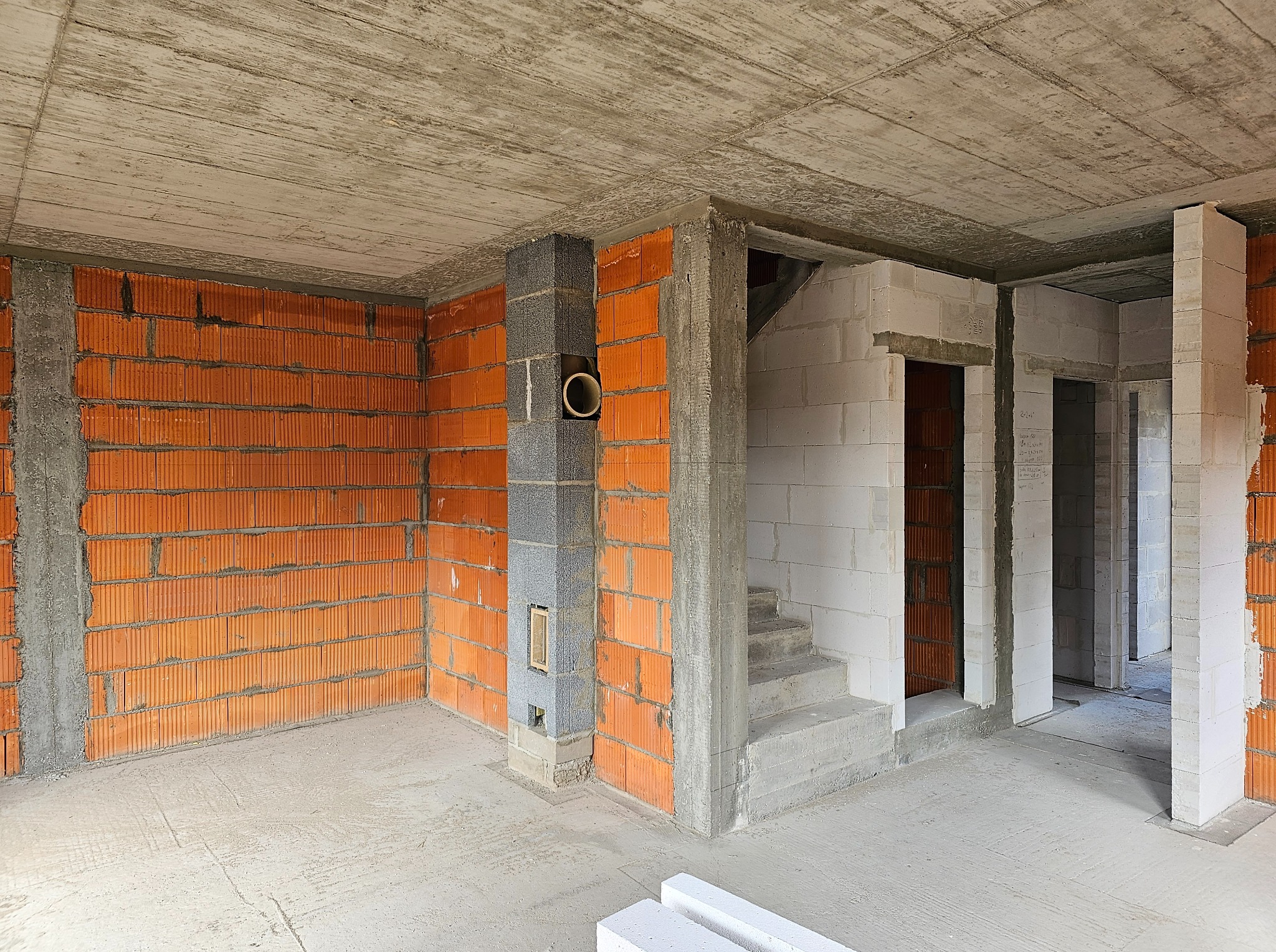 Surowy stan wnętrza domu w trakcie budowy: ściany z czerwonej cegły i białych bloczków, betonowy strop, komin z otworem wentylacyjnym i surowe schody.