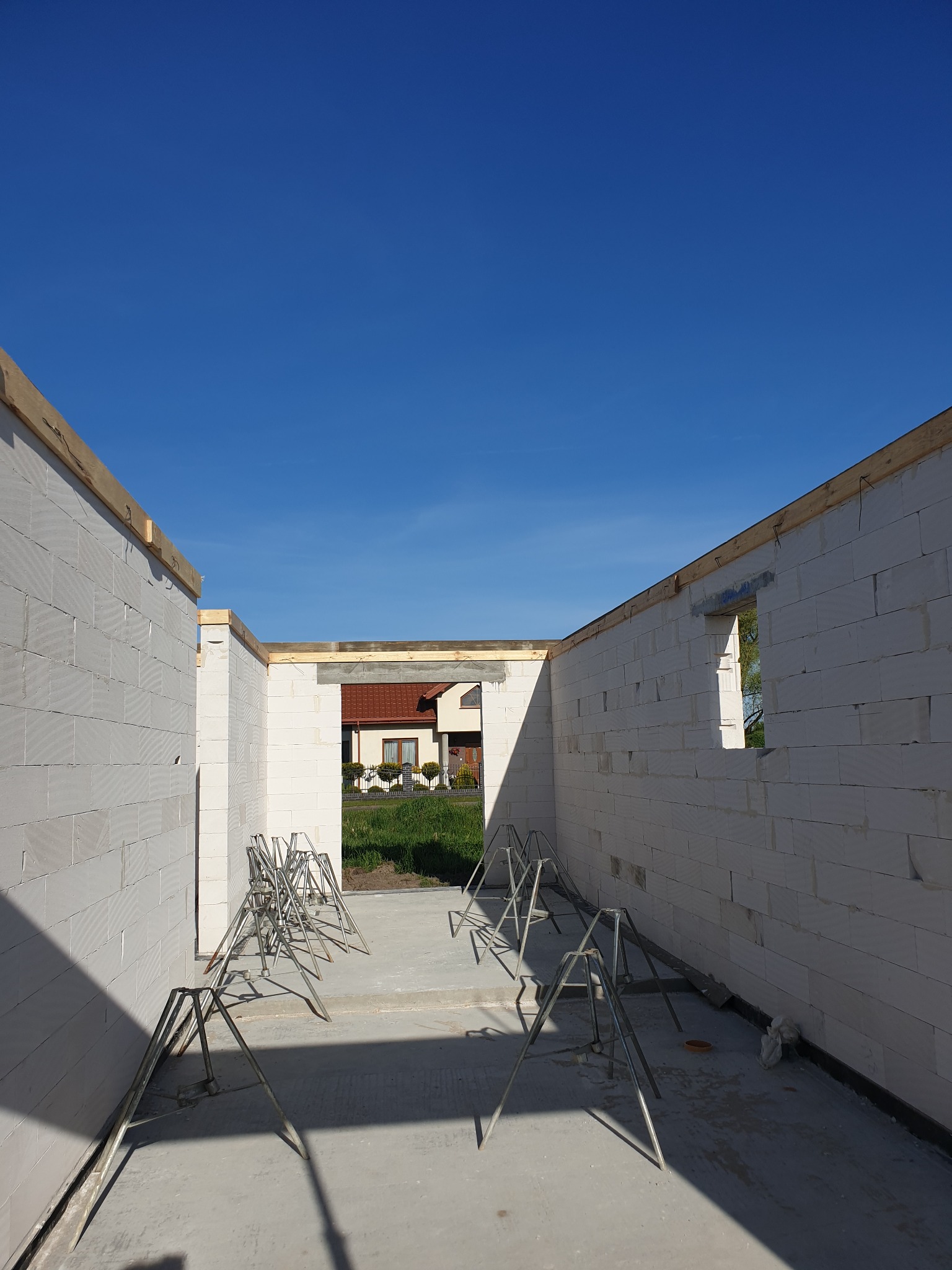 Wnętrze domu murowanego w trakcie budowy, widoczne ściany z bloczków, podpory szalunkowe i fundament wylany betonem; w tle dom z czerwonym dachem.