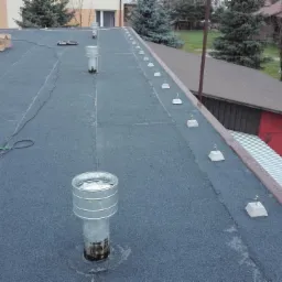 Widok z góry na płaski dach pokryty papą termozgrzewalną z zamontowanymi odpowietrznikami i uchwytami, widoczne są również kartony i przewody, w tle budynki gospodarcze.