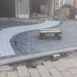 Niedokończona nawierzchnia z szarej i jasnoszarej kostki ułożonej w falisty wzór, widoczny wózek transportowy, wiadro i zagęszczarka w tle.