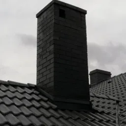 Ciemny, ceglany komin wystający ponad dach pokryty szarą dachówką karpiówką, na tle pochmurnego nieba.