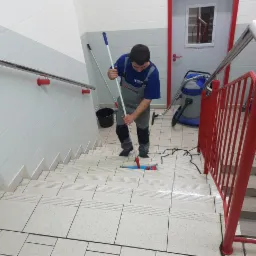 Pracownik w kombinezonie i niebieskiej koszulce myje schody mopem na jasnych płytkach, widoczny odkurzacz przemysłowy, czerwona barierka i biało-szare ściany.