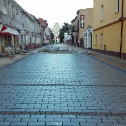 Ulica wyłożona nową, szarą kostką brukową w regularny wzór, z widocznymi paletami z kostką w tle, w perspektywie ulicznej z budynkami po obu stronach.