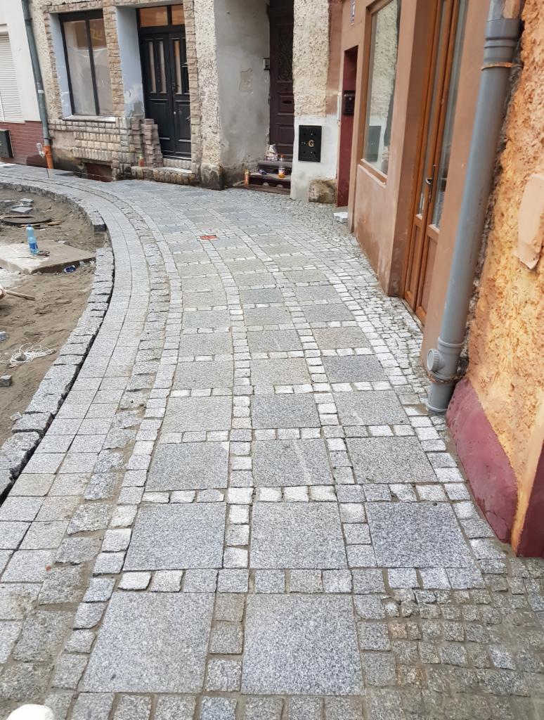 Ułożona ścieżka z kostki granitowej w odcieniach szarości, z widocznymi elementami przygotowania podłoża i otoczeniem budynków.