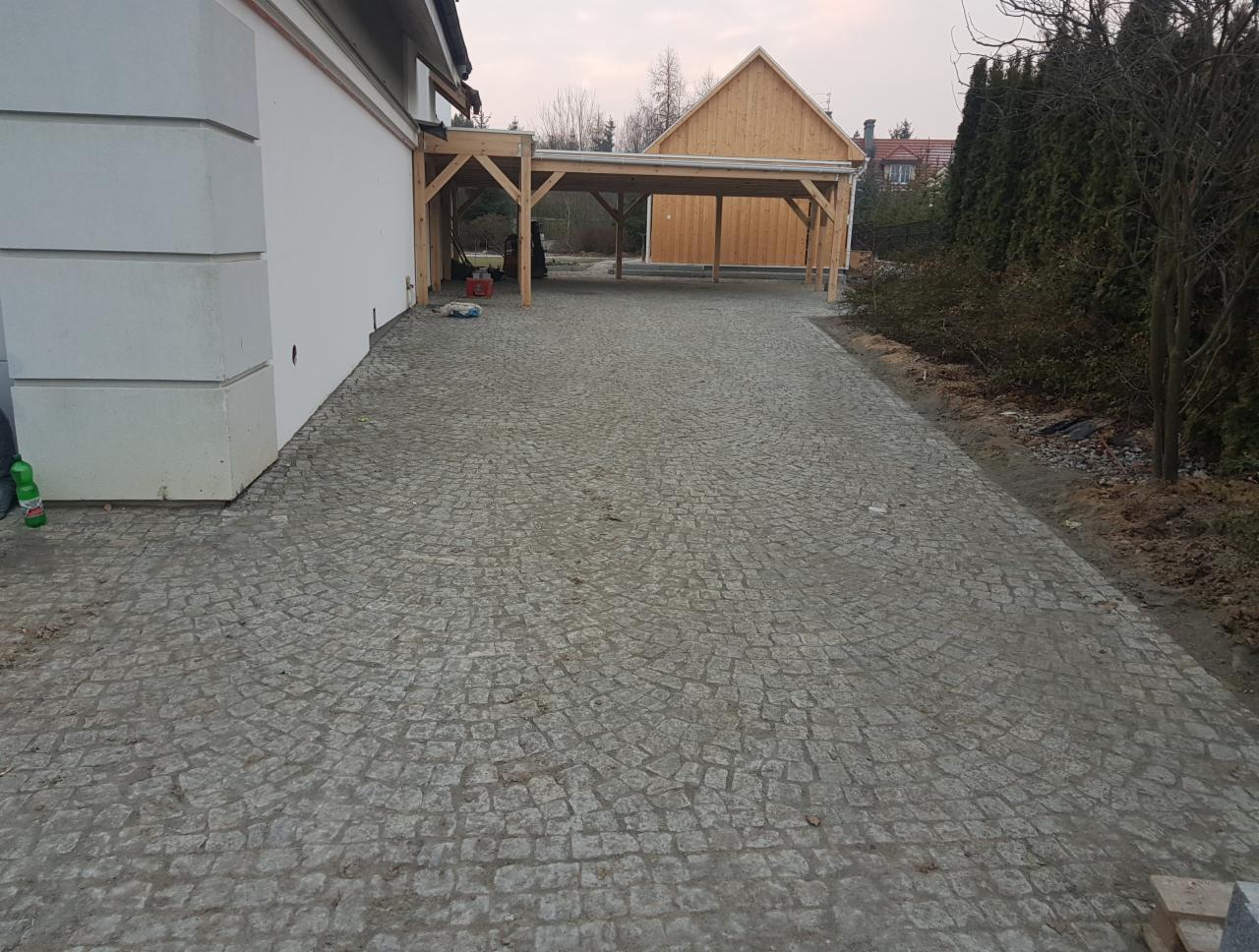 Szeroki, wyłożony szarą kostką plac przed domem z drewnianą wiatą garażową, widok z perspektywy osoby stojącej na placu, w tle iglaki.