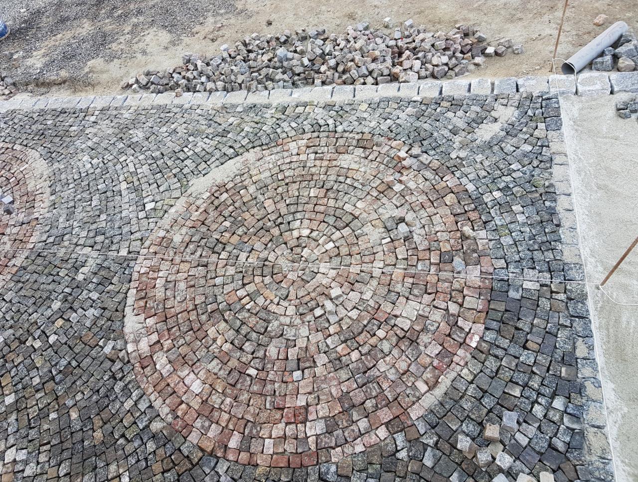 Fragment powierzchni z kostki granitowej ułożonej w okrągły wzór, z widocznym zapasem kostki i rurą PCV w tle.