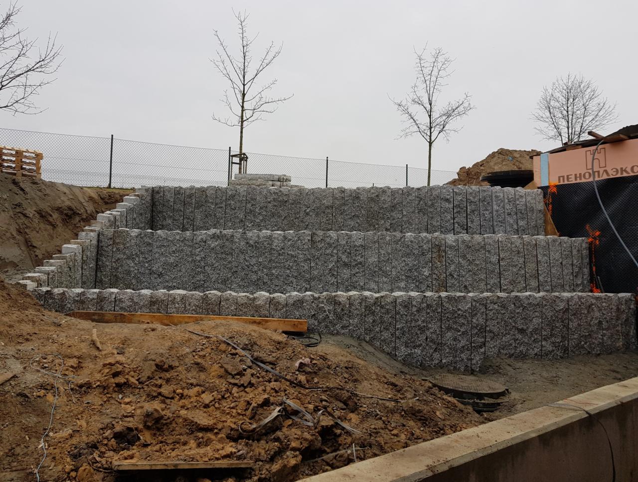 Trzy poziomy tarasów z kostki granitowej w trakcie budowy, widoczne schody z kostki, ziemia, materiały izolacyjne i drzewa w tle.