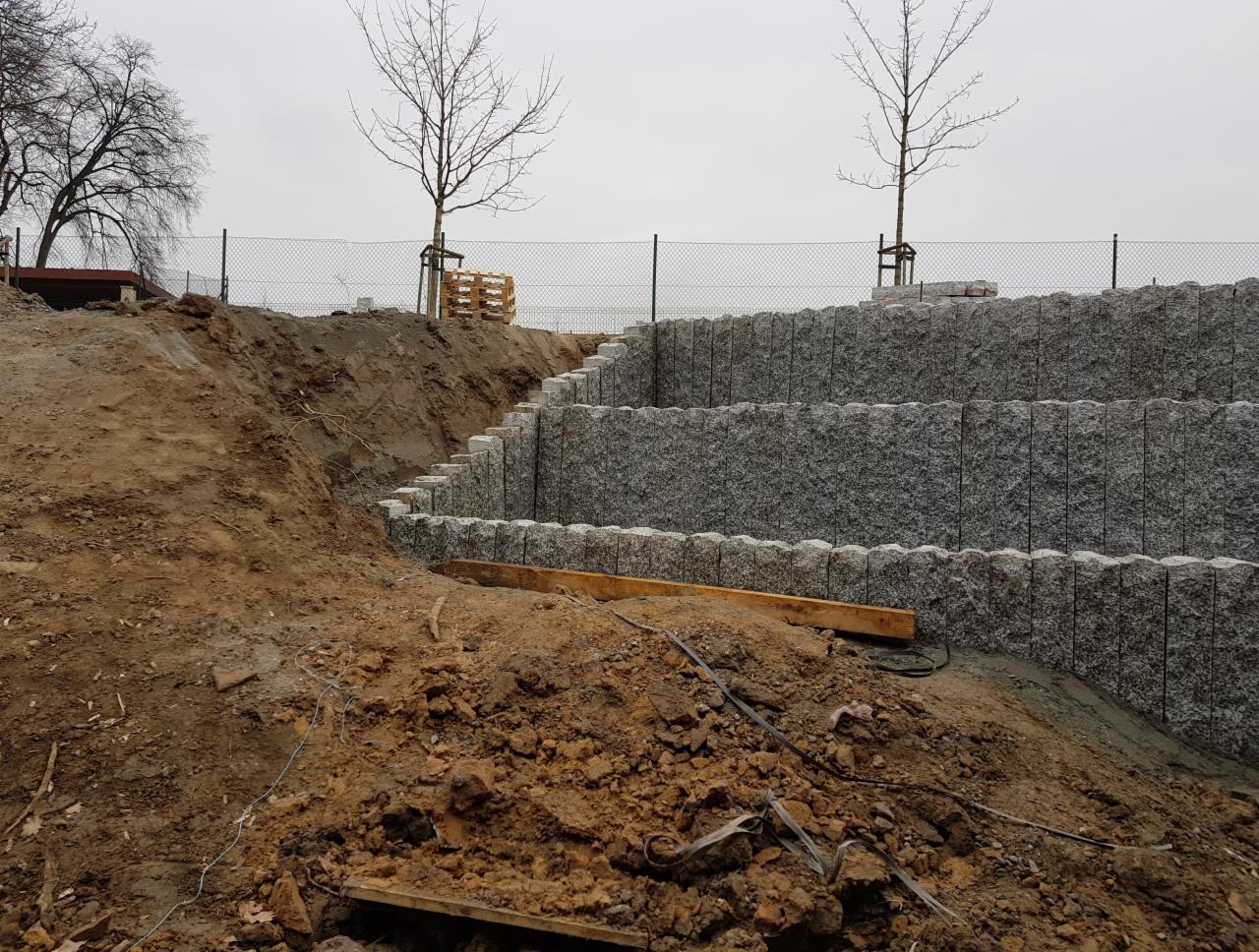Budowa tarasowego muru oporowego z szarej kostki granitowej, widoczne etapy prac ziemnych i ułożone elementy, tło z drzewami bez liści i siatką ogrodzeniową.