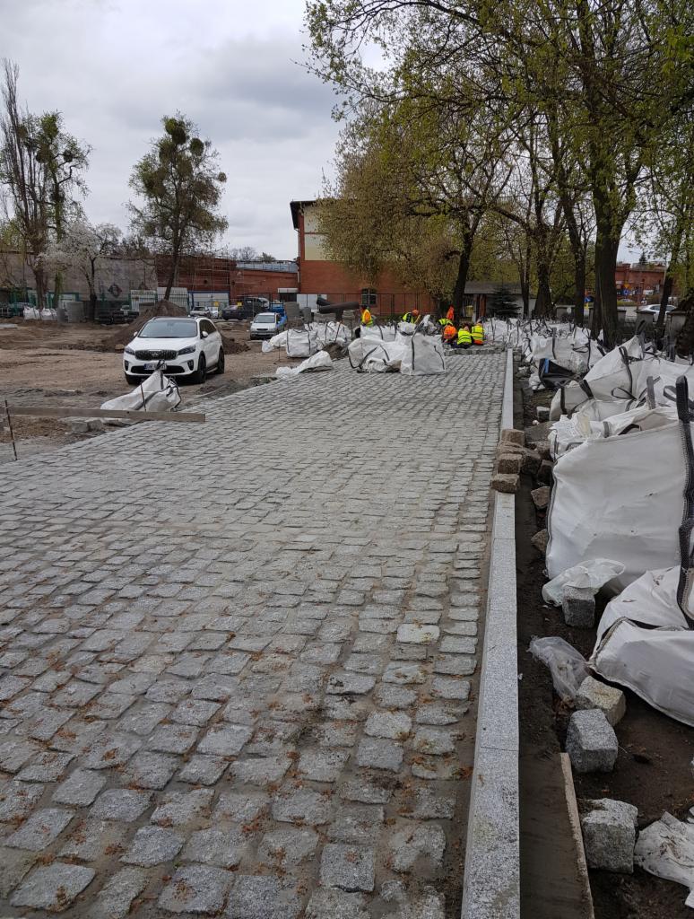 Świeżo ułożona nawierzchnia z kostki granitowej w odcieniach szarości, z widocznymi pracownikami w odblaskowych kamizelkach i workami z materiałem w tle, podczas dnia.