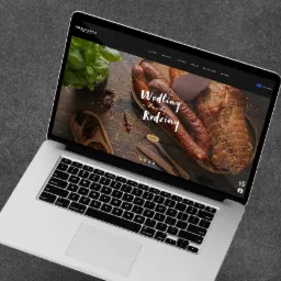 Laptop wyświetla stronę internetową z wędlinami na drewnianym talerzu, z ziołami w tle, prezentowaną jako 'Wędliny dla całej rodziny'.