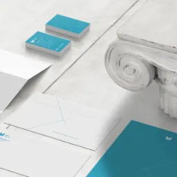 Komplet materiałów firmowych: wizytówki, papier firmowy, koperty i teczka w kolorach bieli i turkusu, ułożone obok detalu architektonicznego kolumny na białym drewnianym stole.