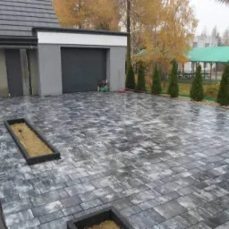 Ułożona szara kostka brukowa prowadząca do garażu nowoczesnego domu, z widocznymi rabatami kwiatowymi wbudowanymi w nawierzchnię i elementami w trakcie wykończenia.