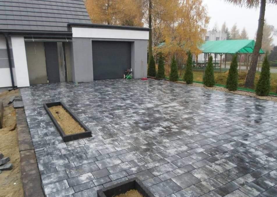 Ułożona szara kostka brukowa prowadząca do garażu nowoczesnego domu, z widocznymi rabatami kwiatowymi wbudowanymi w nawierzchnię i elementami w trakcie wykończenia.