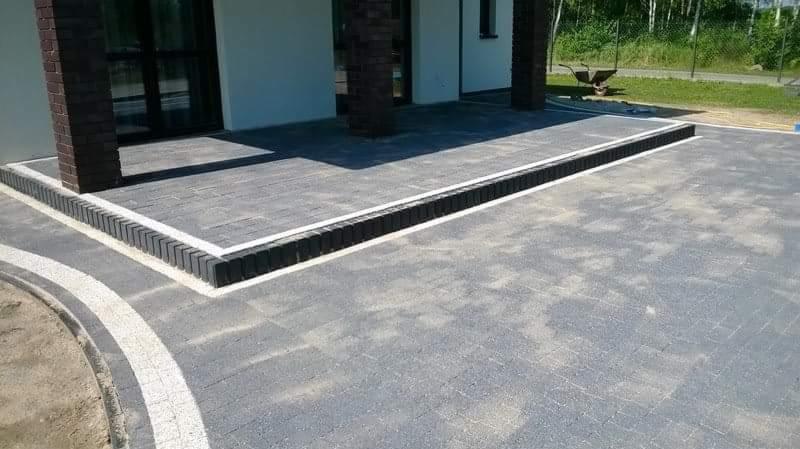 Ułożona kostka brukowa w odcieniach szarości z dekoracyjnym obramowaniem z ciemnej kostki, widok na fragment elewacji domu z ceglanymi filarami i wózkiem budowlanym w tle.