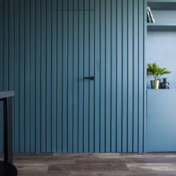 Niebieska ściana z ukrytymi drzwiami z pionowymi panelami, w tle zabudowa meblowa z półkami i szafkami, minimalistyczny design.