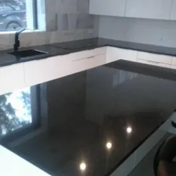 Nowoczesna kuchnia z czarnym blatem, białymi szafkami i widokiem z okna odbijającym się w powierzchni blatu.