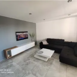 Nowoczesny salon z narożną sofą w kolorze antracytu, białą szafką RTV z drewnianymi akcentami, stolikiem kawowym z białych płyt i szarą ścianą z telewizorem.