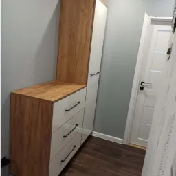 Wysoka szafka z szufladami i p&oacute;łką z drewnopodobnym wykończeniem i białymi frontami, czarne uchwyty, ustawiona w rogu pokoju z panelami podłogowymi.