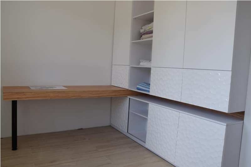 Białe szafy z dekoracyjnymi frontami i drewniany blat biurka na czarnej nodze, minimalistyczne wnętrze.