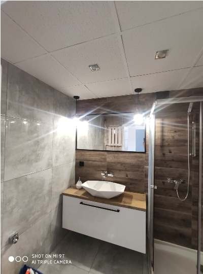 Nowoczesna łazienka z umywalką nablatową w kształcie diamentu, szafką z drewnianym blatem i czarnym uchwytem, lustrem w czarnej ramie i kabiną prysznicową z drewnopodobnymi płytkami.