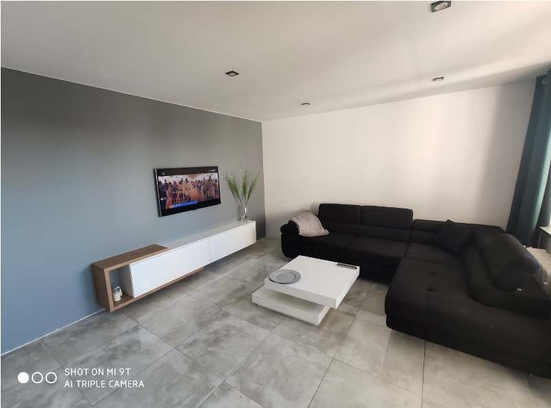 Nowoczesny salon z narożną sofą w kolorze antracytu, białą szafką RTV z drewnianymi akcentami, stolikiem kawowym z białych płyt i szarą ścianą z telewizorem.