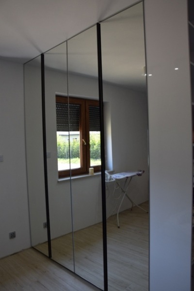 Wysoka szafa z lustrzanymi drzwiami przesuwnymi w Częstochowie, z czarnymi ramami, odbija okno z roletami i deskę do prasowania.