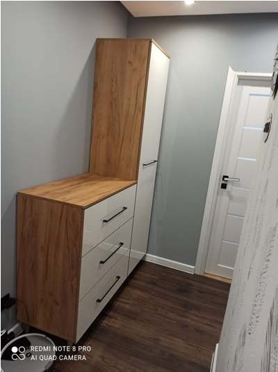 Wysoka szafka z szufladami i półką z drewnopodobnym wykończeniem i białymi frontami, czarne uchwyty, ustawiona w rogu pokoju z panelami podłogowymi.