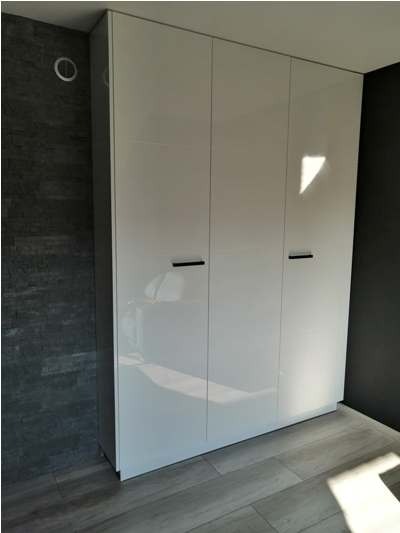 Trzydrzwiowa, biała szafa wnękowa z połyskiem, z minimalistycznymi, czarnymi uchwytami, na tle ściany wykończonej płytkami imitującymi kamień i podłogi z paneli w odcieniach szarości.