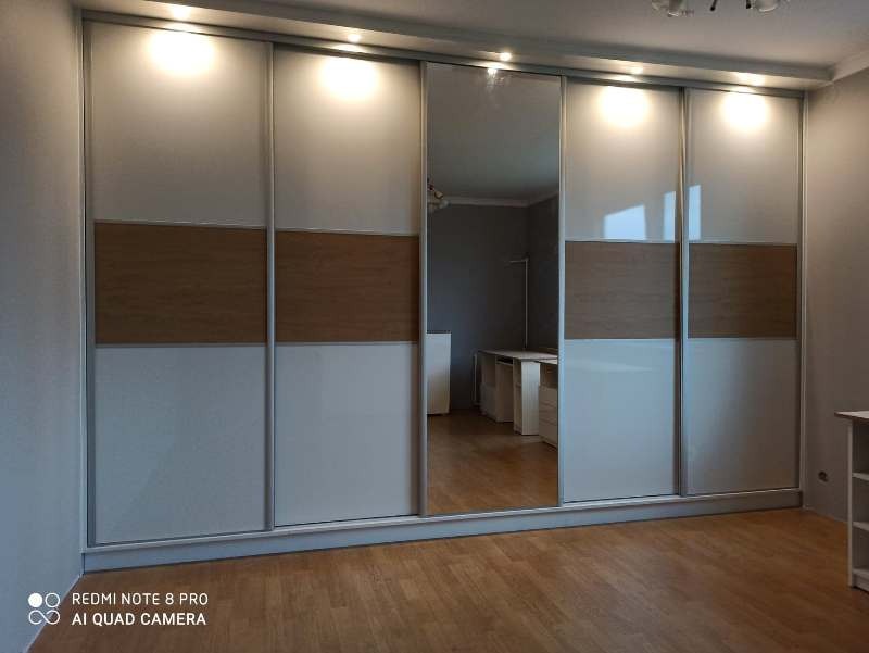 Duża szafa przesuwna z lustrzanymi drzwiami i drewnianym akcentem, oświetlona od góry, widoczna w przestronnym wnętrzu z parkietem.