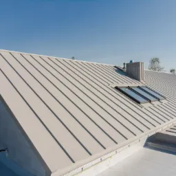 Widok z góry na jasnoszary dach z blachy z trzema oknami dachowymi i kominem, na tle błękitnego nieba.