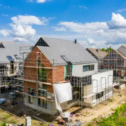 Wysoka perspektywa na kilka domów w trakcie budowy z szarą blachą trapezową na dachach, widoczne rusztowania, ceglane elewacje i instalacja okien dachowych, w tle błękitne niebo z chmurami i żuraw...