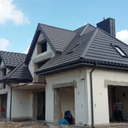 Dom w trakcie budowy z nowym, ciemnoszarym dachem z dachówki ceramicznej, rynnami w kolorze dachu, oknem dachowym i kominem z blachy. Widoczny pracownik w garażu.