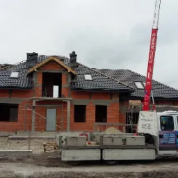 Budowa domu jednorodzinnego w trakcie, widoczny mur z czerwonej cegły, częściowo położone czarne pokrycie dachowe, okna dachowe, komin z cegły klinkierowej, rusztowania, podnośnik dekarski...