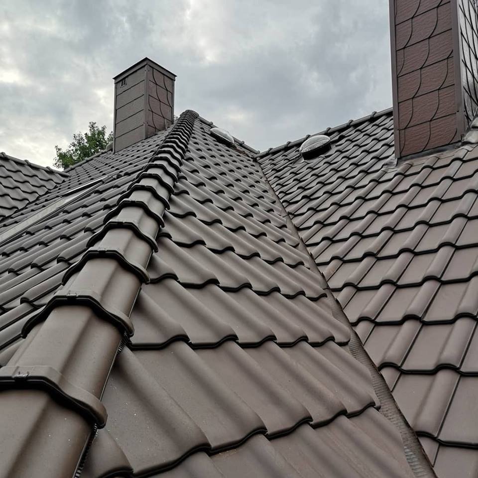 Brunatny dach z dachówki ceramicznej z dwoma kominami i świetlikami dachowymi, widziany pod kątem w pochmurny dzień.
