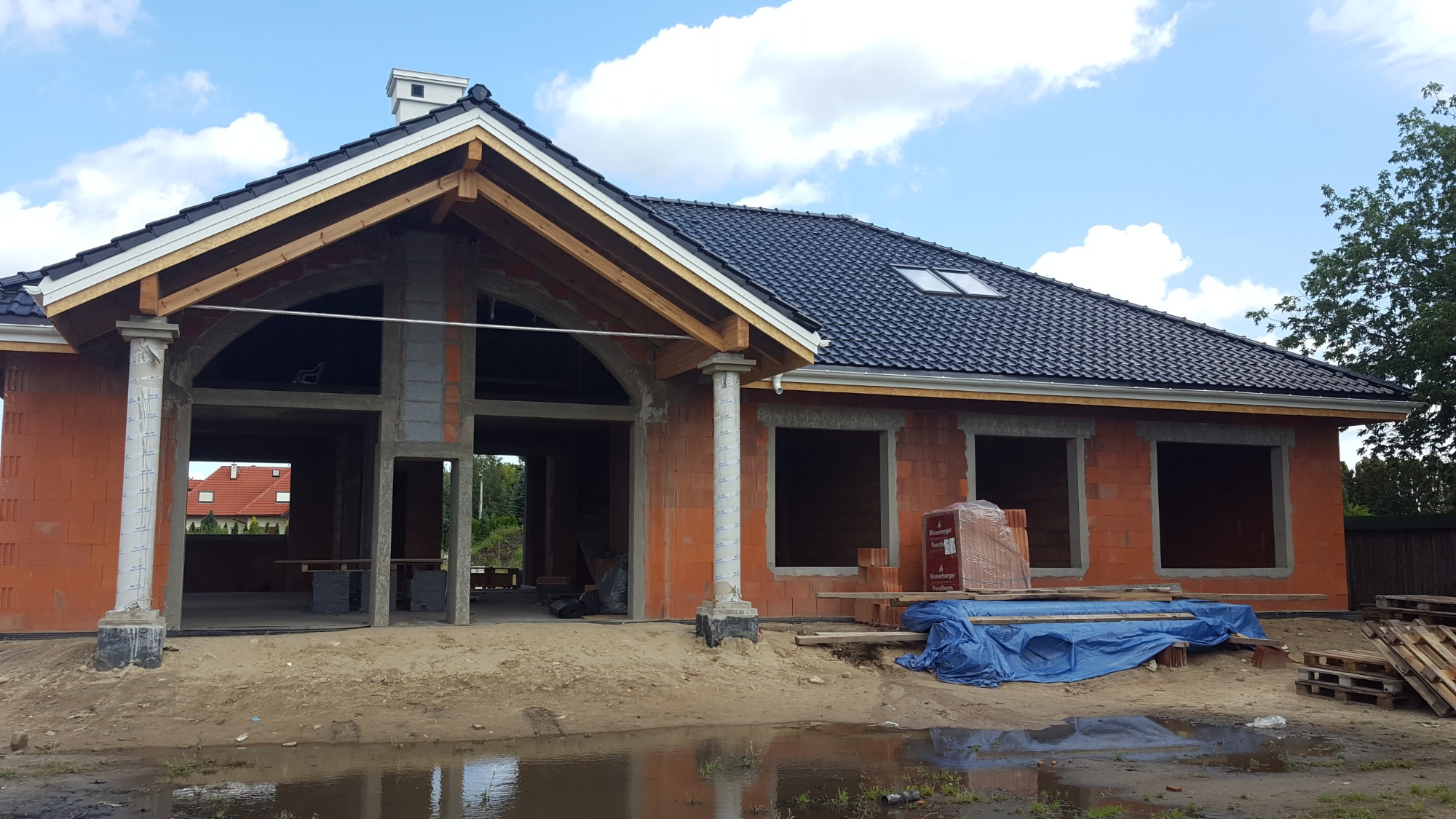 Dom w trakcie budowy z widoczną konstrukcją dachu pokrytą czarną dachówką, z elementami więźby dachowej i widocznymi rynnami, ściany z czerwonej cegły, palety z cegłami pod plandeką.
