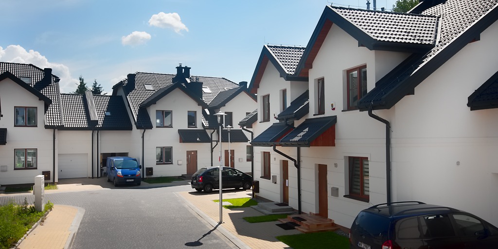 Nowoczesne szeregowe domy z ciemną dachówką i jasnymi elewacjami, widok z ulicy osiedlowej, samochody zaparkowane przed budynkami.