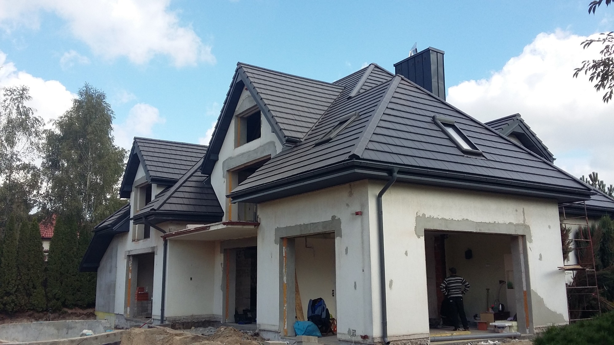 Dom w trakcie budowy z nowym, ciemnoszarym dachem z dachówki ceramicznej, rynnami w kolorze dachu, oknem dachowym i kominem z blachy. Widoczny pracownik w garażu.