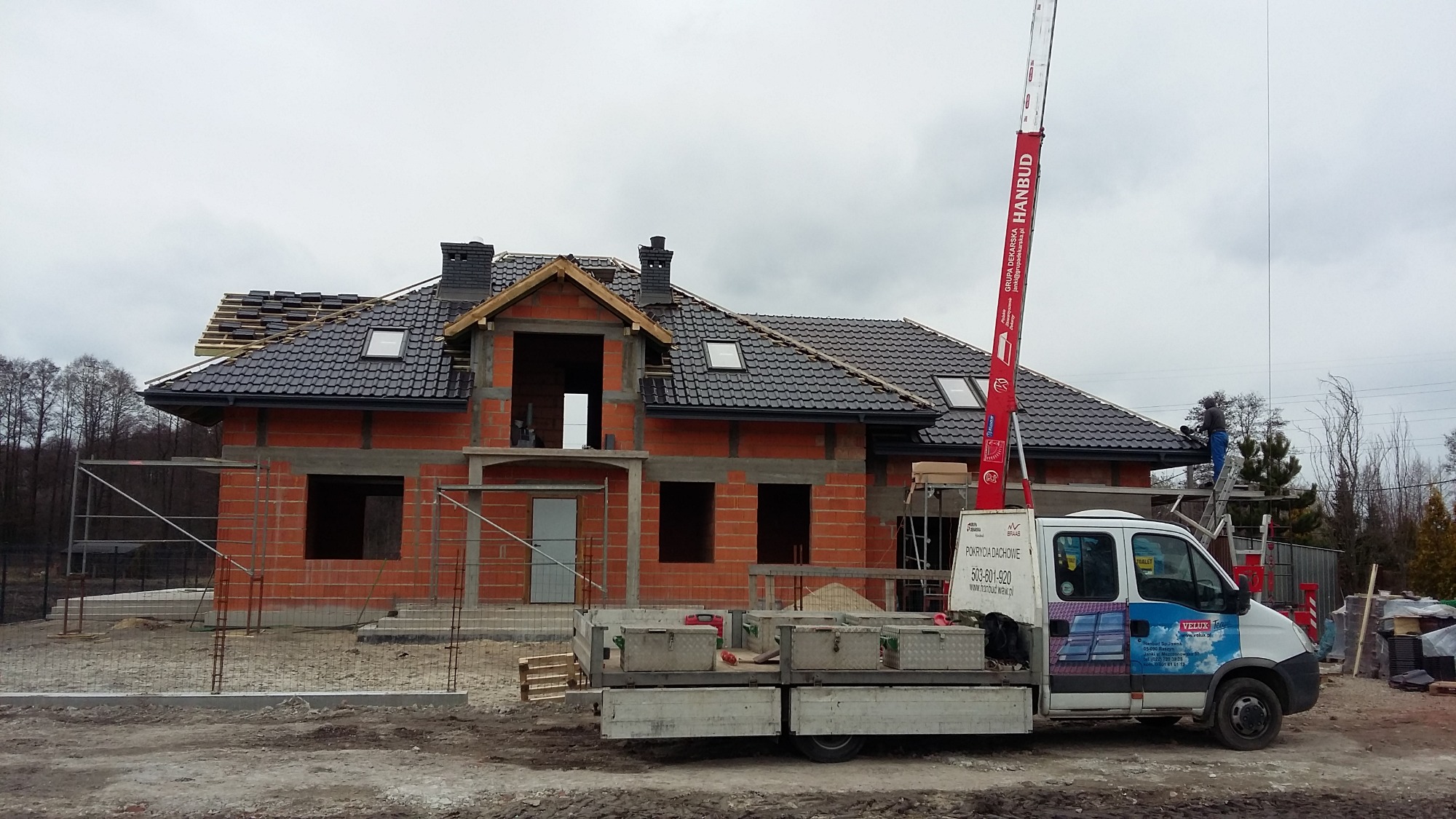 Budowa domu jednorodzinnego w trakcie, widoczny mur z czerwonej cegły, częściowo położone czarne pokrycie dachowe, okna dachowe, komin z cegły klinkierowej, rusztowania, podnośnik dekarski...