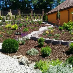 Ukształtowany, wielopoziomowy ogród z krętą ścieżką z białego żwiru, obrzeżami z betonowych elementów oraz roślinami okrywowymi i ozdobnymi krzewami, z budynkiem w tle.