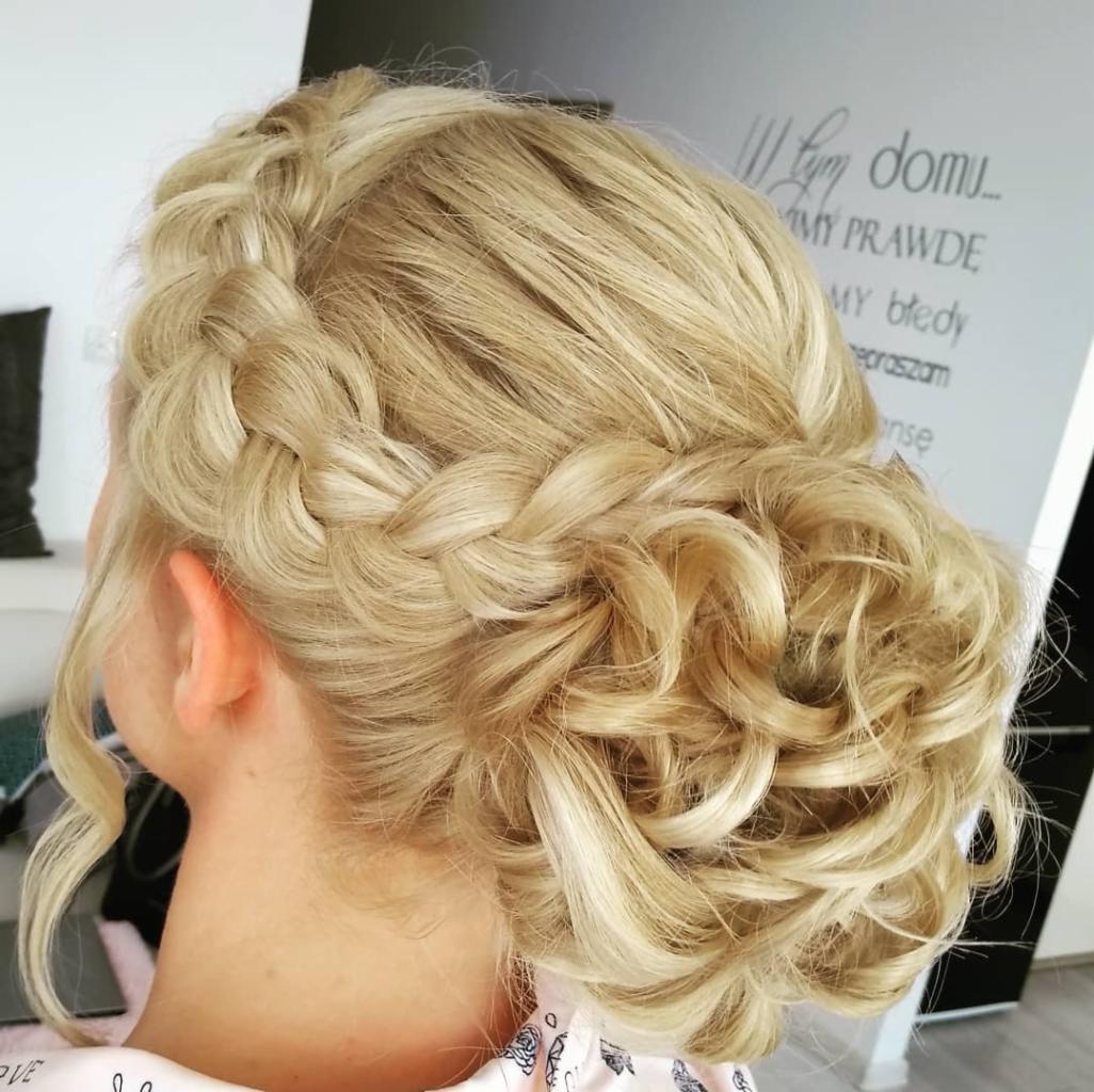 Upięcie blond włosów z warkoczem i lokami, widok z tyłu, subtelne oświetlenie, tekst na ścianie w tle.