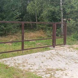 Arek - Brązowa, dwuskrzydłowa brama z siatki panelowej na posesji z betonową kostką, widok na las w tle.