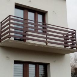 Arek - Balkon z brązową metalową balustradą z poziomych profili, widok z dołu na tle białej elewacji i pochmurnego nieba.