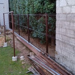 Arek - Montaż brązowego ogrodzenia z paneli siatkowych na betonowej podmurówce, widoczne słupki i elementy montażowe, w tle wysokie tuje i budynek z bloczków betonowych.